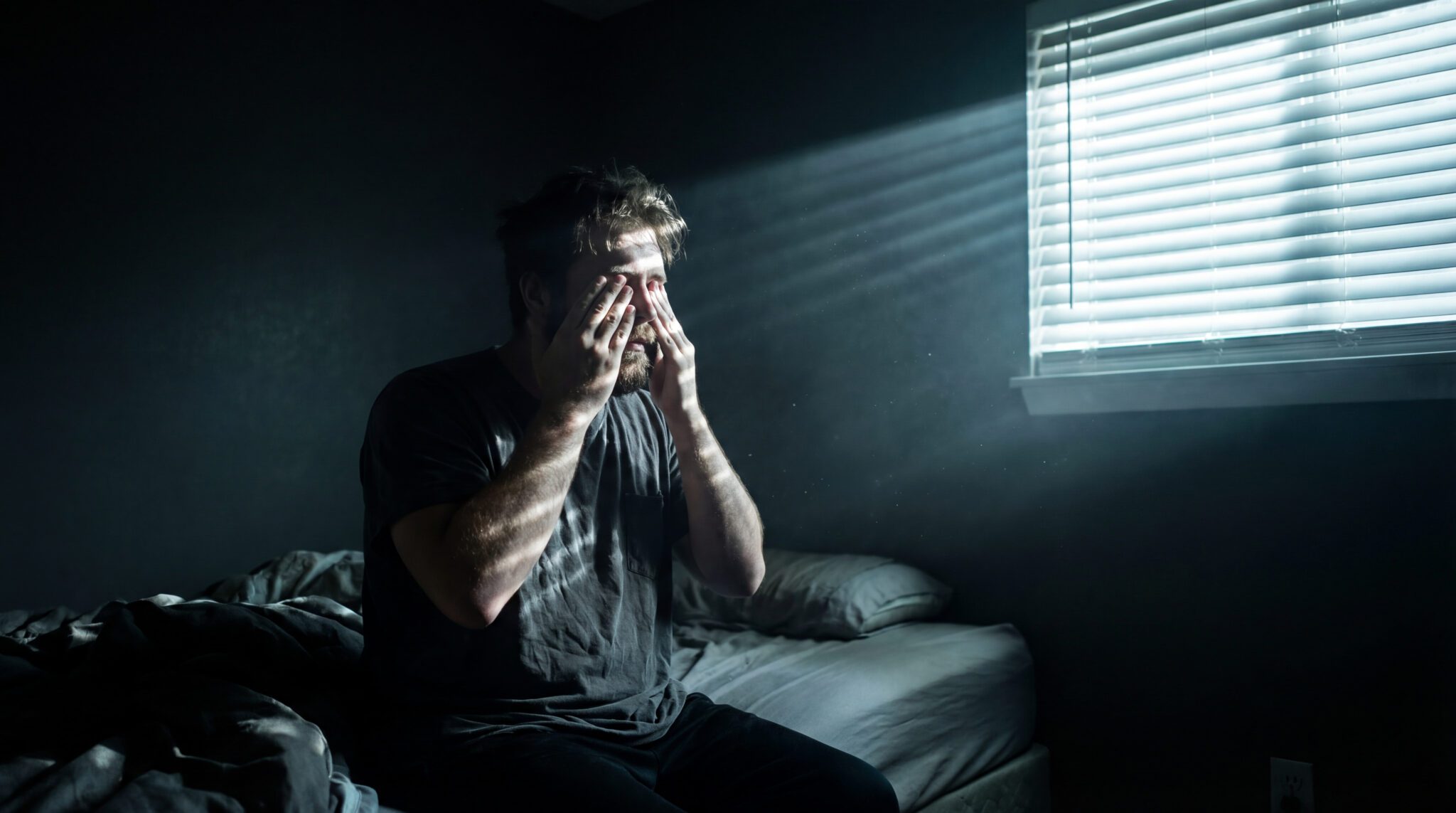 Ein übermüdeter Mann sitzt nachts auf der Bettkante, während grelles LED-Außenlicht durch die Jalousien in das dunkle Schlafzimmer strahlt.