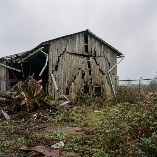 Die massiv beschädigte, rissige Holzgiebelwand eines ruinösen Schuppens auf einem verwahrlosten Gewerbegelände.