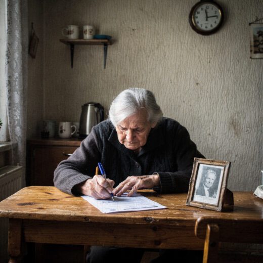 Eine ältere Frau fälscht angespannt am Tisch eine Unterschrift auf einem Brief; daneben liegt ein Foto ihres verstorbenen Partners.