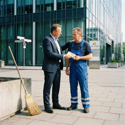 Ein Mann im Anzug übergibt einem Arbeiter vor einer Glasfront vertraulich einen dicken Stapel Geldscheine.